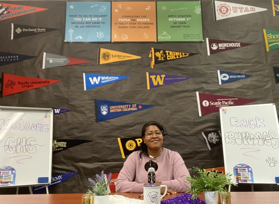 Ms. Tiffany sitting at a table with a podcast microphone. A collage of various college banners on the wall behind Ms. Tiffany